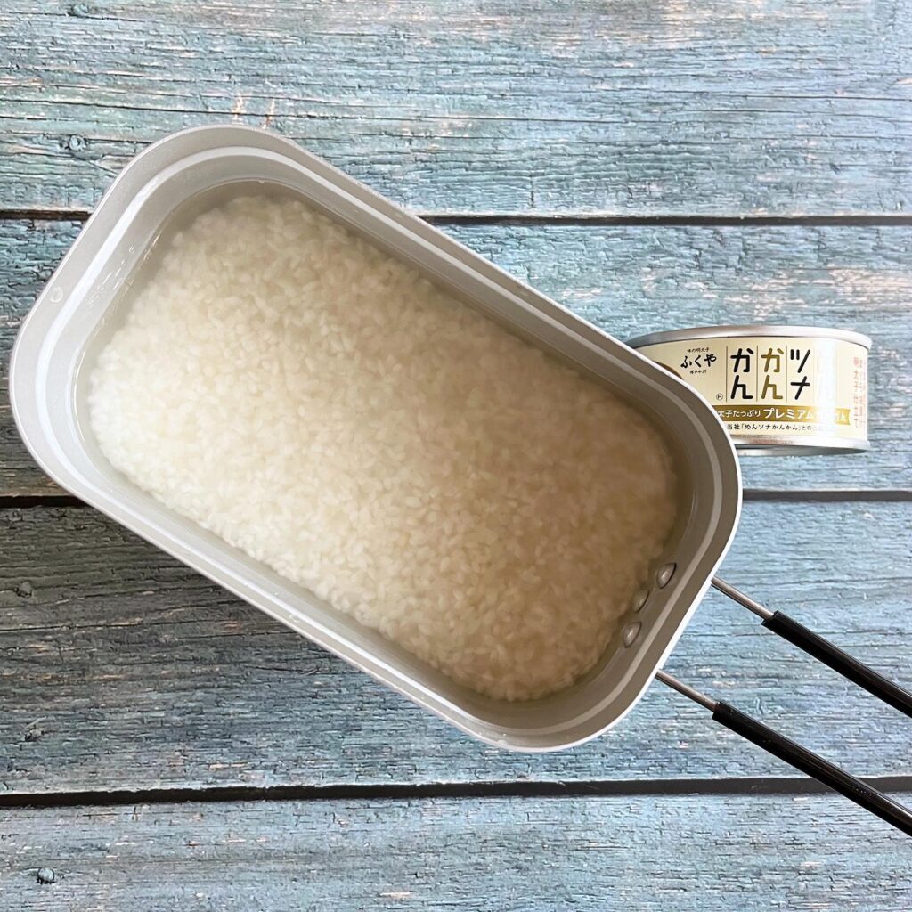 メスティンにお米と水がいれてあり、そのそばにめんツナかんかんが置いてある調理前の写真